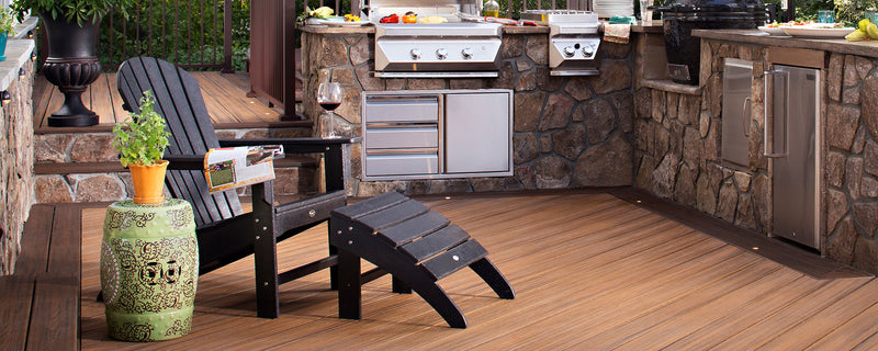 A black Trex Outdoor Furniture Cape Cod Adirondack chair and ottoman sit near an outdoor kitchen on a brown composite deck.