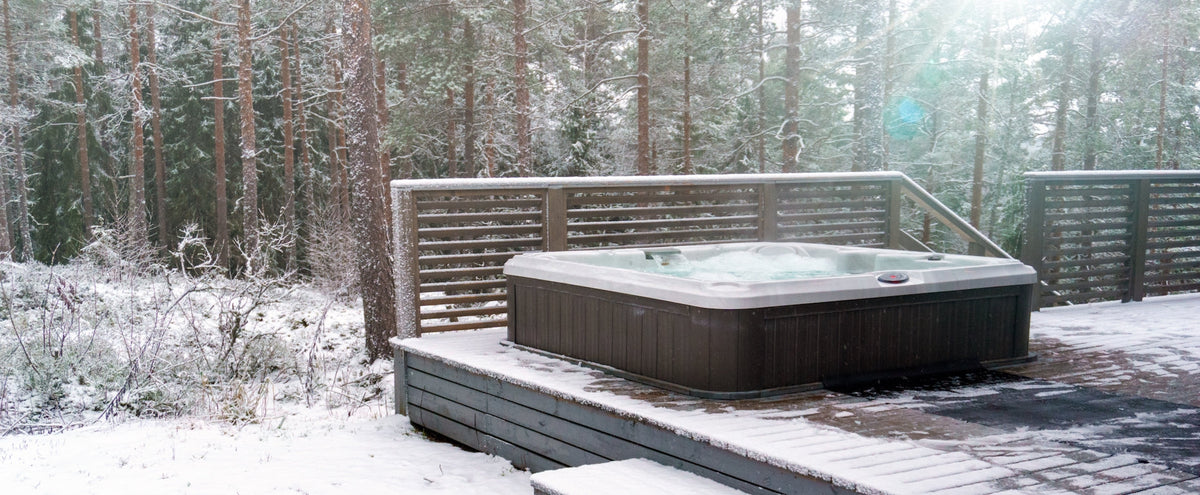 In a snowy winter scene, steam rises from the churning water of a hot tub on a deck next to a thick forest.