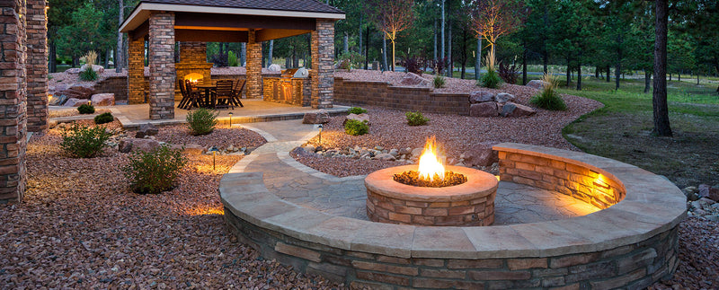 A stone patio features an outdoor kitchen, a round fire pit area, and landscaping with lava rocks, lighting, and shrubs.