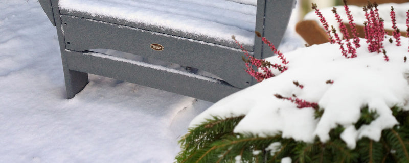 A grey Trex Outdoor Furniture Adirondack chair is covered in snow near an evergreen bush.