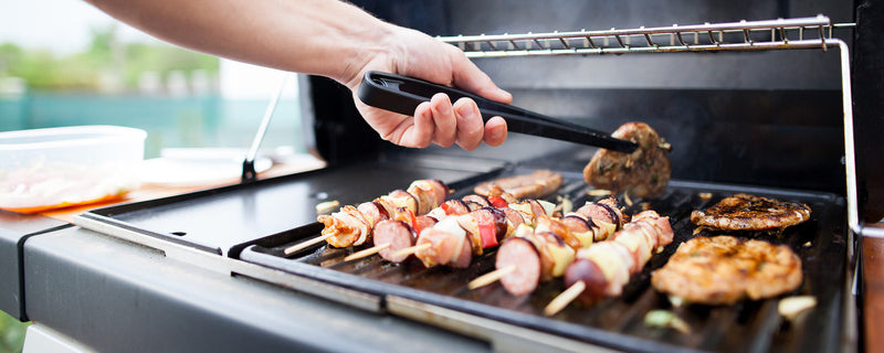 A hand uses tongs to flip chicken breasts on a grill behind skewers of sausage and vegetables.