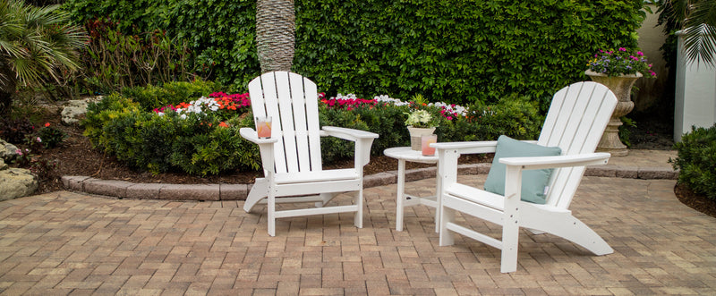 A white Trex Outdoor Furniture Yacht Club 3 Piece Adirondack Set sits on a curved brick patio before flowers and shrubs.