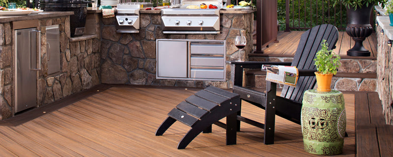 A black Trex Outdoor Furniture Yacht Club Adirondack Chair and ottoman sit by a stone outdoor kitchen on a brown deck.