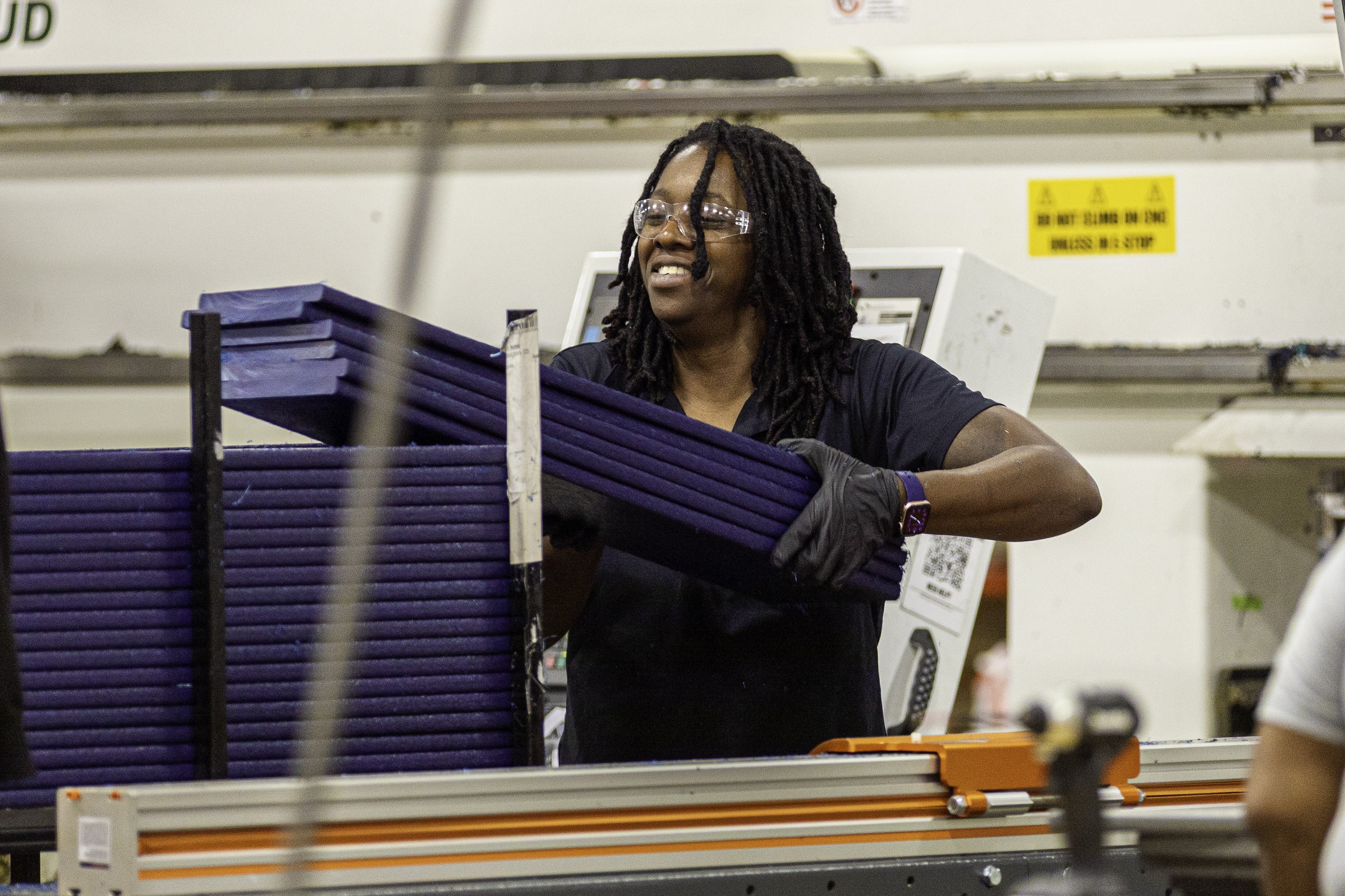 A person wearing safety goggles and gloves stacks navy blue HDPE lumber planks in a factory setting.