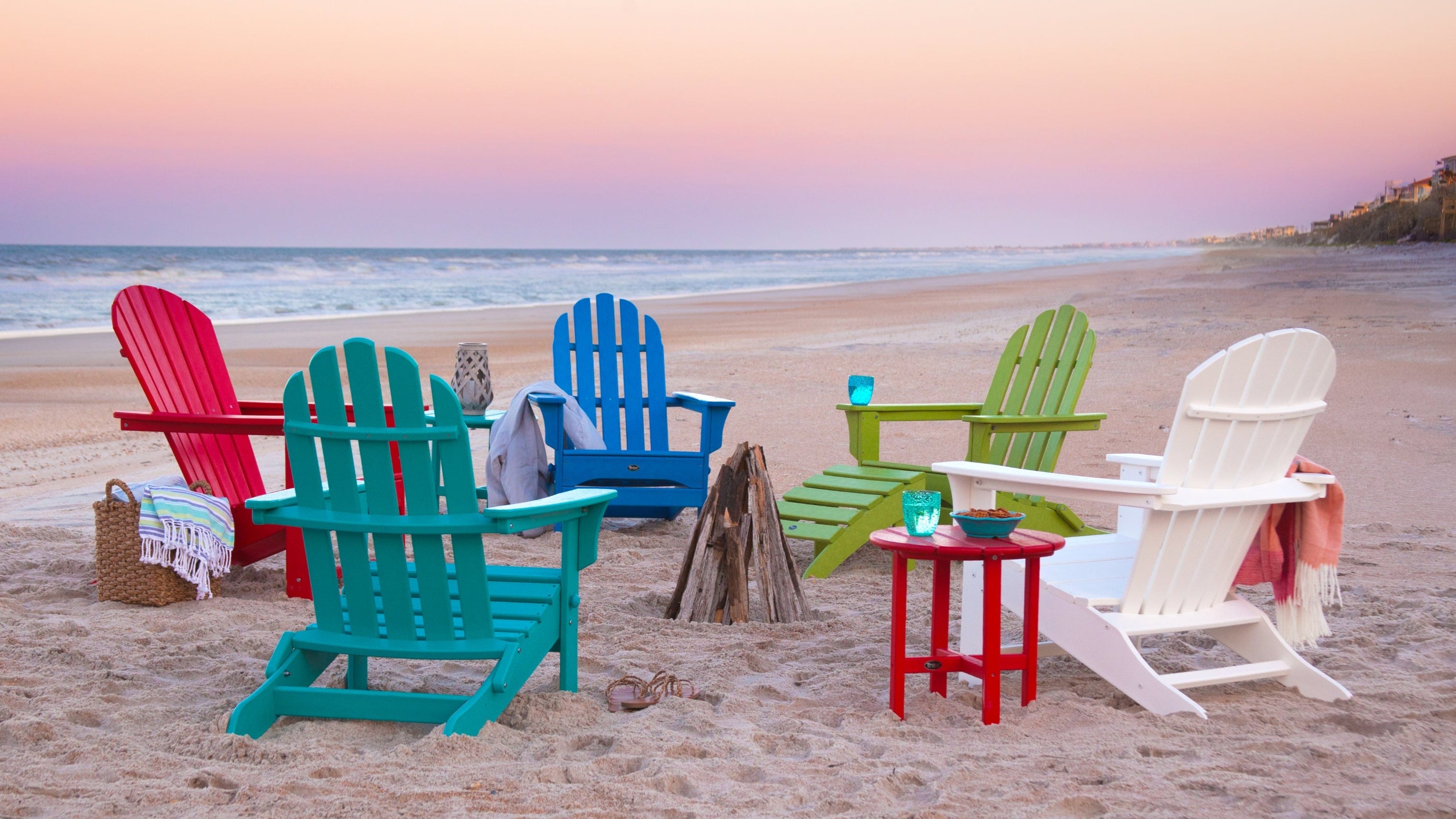 Colorful Trex Outdoor Furniture Adirondack chairs sit on a sandy beach with a sunset sky.
