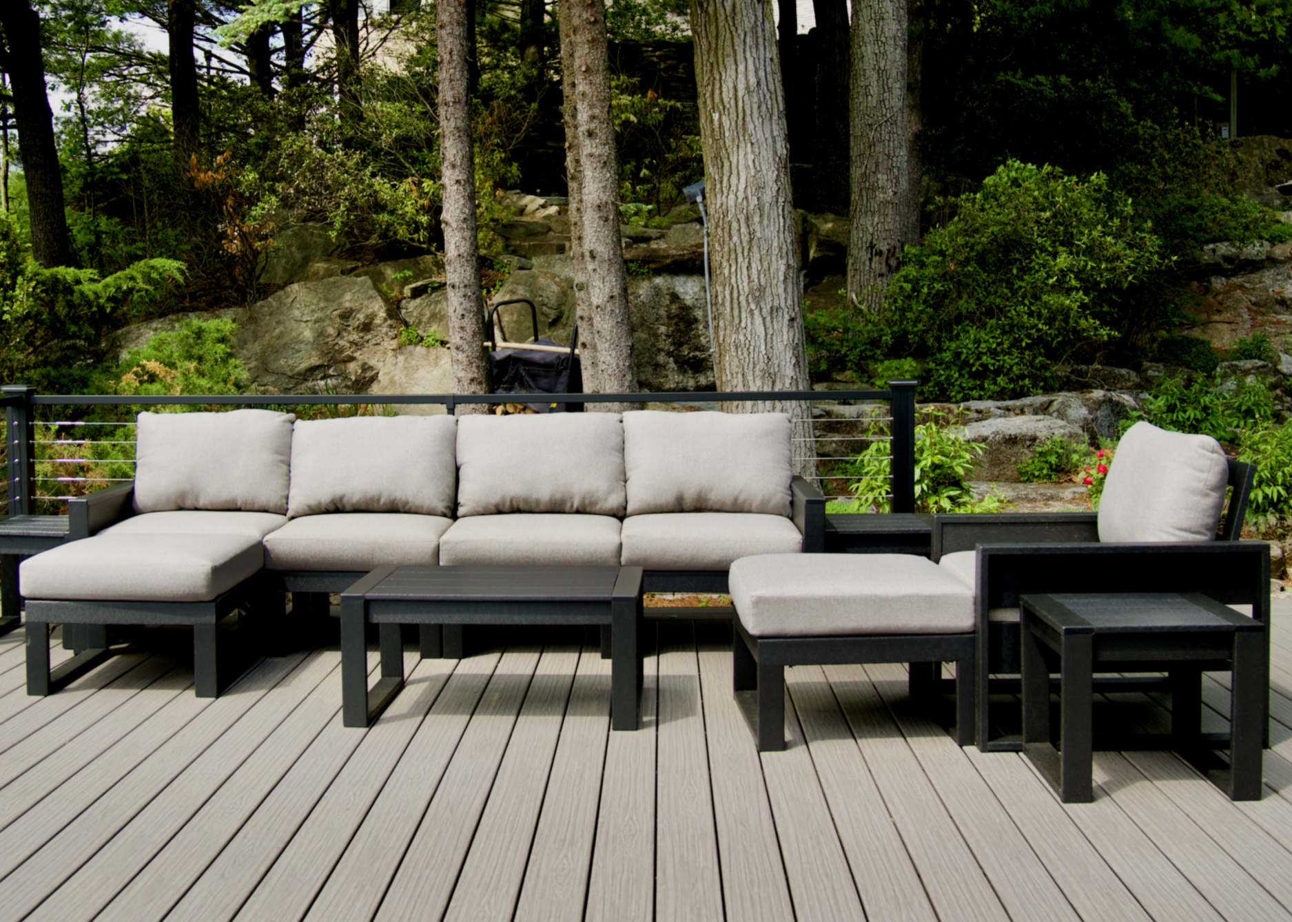 A black Trex Outdoor Furniture Eastport sectional with cream cushions sits on a wooden deck surrounded by trees.