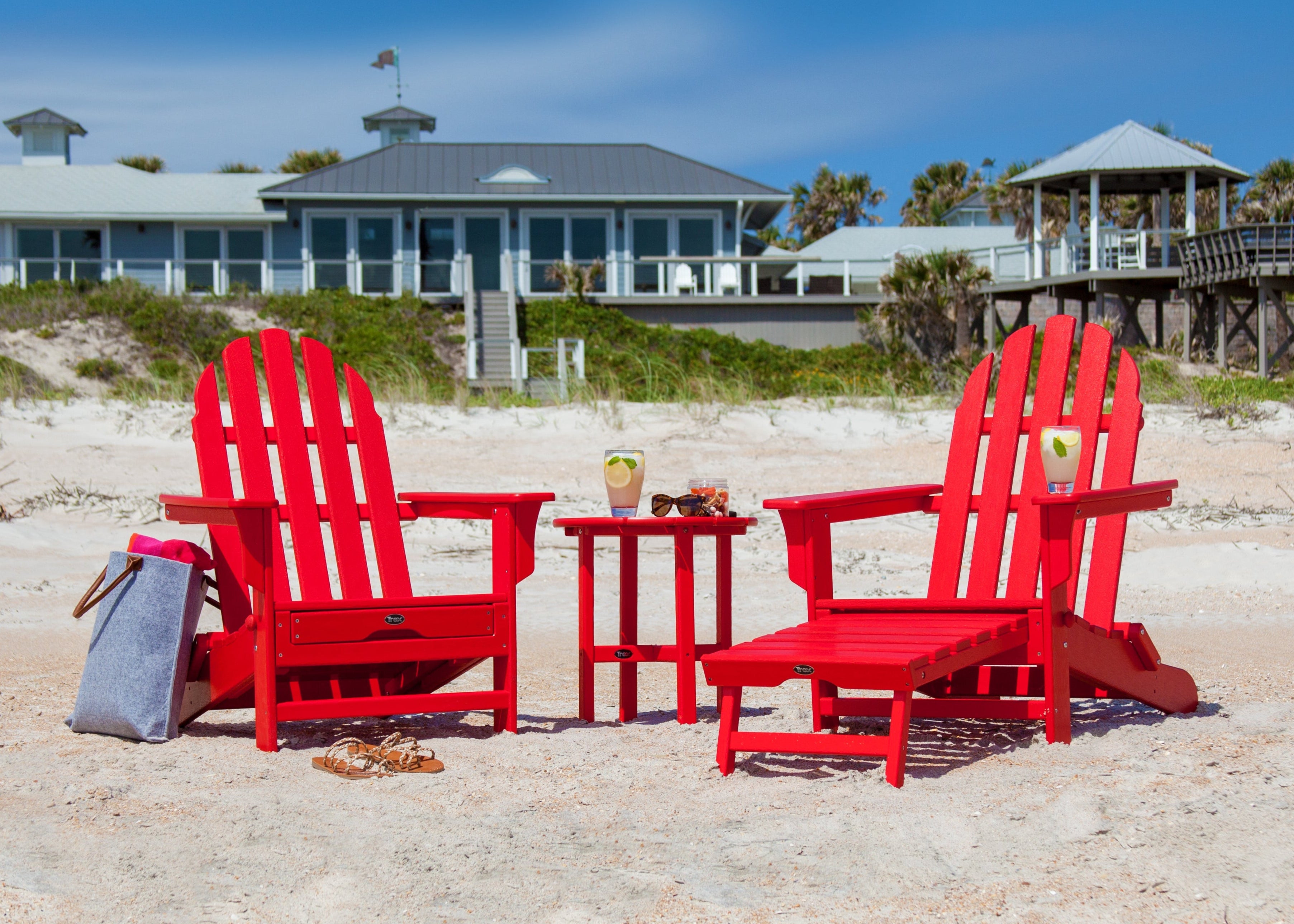 Two red Trex Outdoor Furniture Cape Cod Ultimate Adirondack chairs and a small table sit on a sandy beach with buildings in the background.