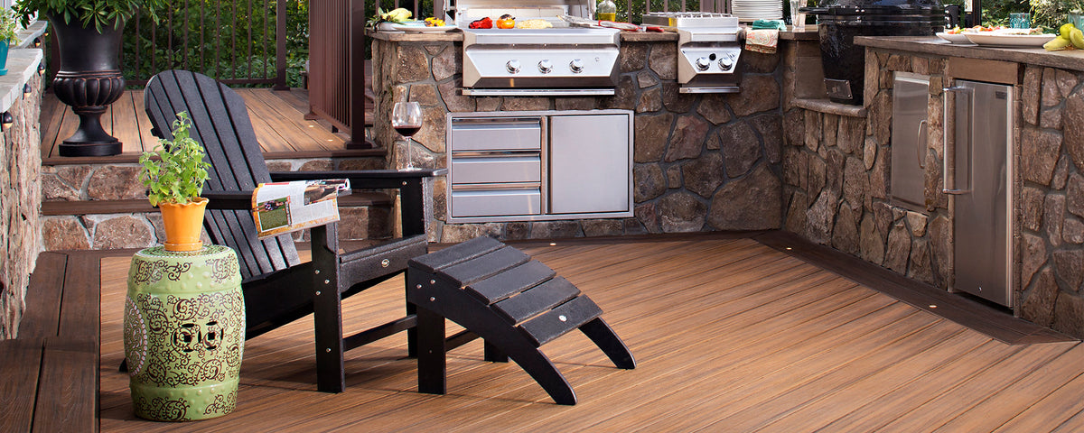 A black Trex Outdoor Furniture Cape Cod Adirondack chair and ottoman sit near an outdoor kitchen on a brown composite deck.