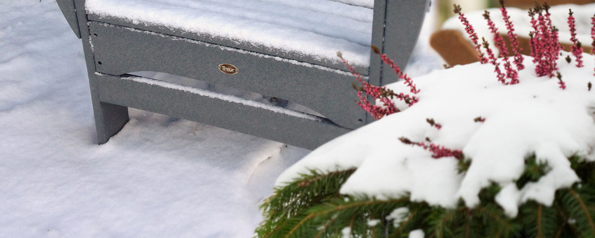 A grey Trex Outdoor Furniture Adirondack chair is covered in snow near an evergreen bush.