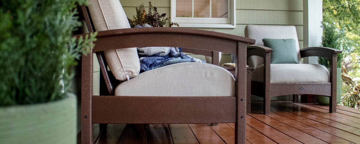 Two brown Trex Outdoor Furniture Rockport Club Deep Seating Chairs with beige cushions sit on a green house's wooden porch.