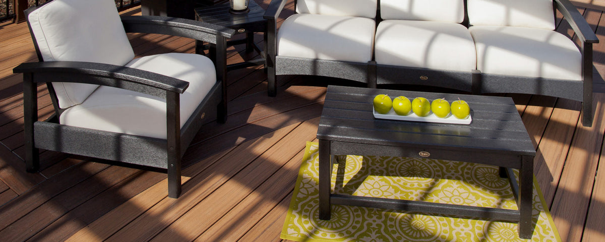 A black Trex Outdoor Furniture Rockport coffee table and a deep seating chair and sofa with white cushions sit on a deck.