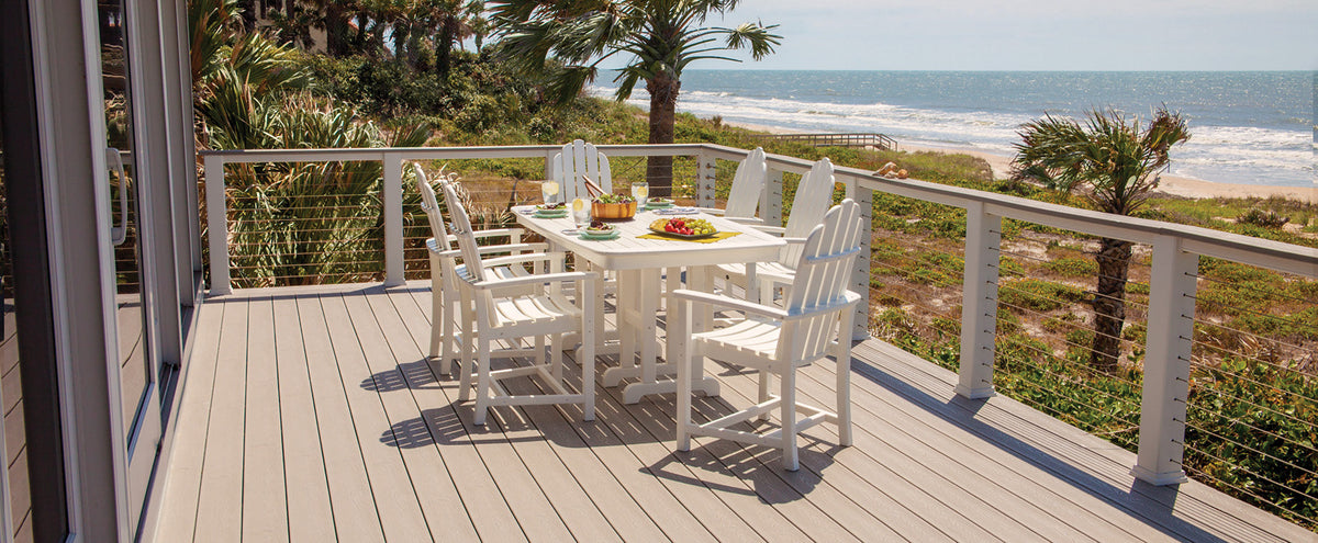 A white Trex Outdoor Furniture Cape Cod Adirondack 7 Piece Dining Set sits on a deck overlooking a beach and the ocean.