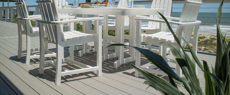 A white Trex Outdoor Furniture Cape Cod Adirondack 7 Piece Dining Set sits on a deck overlooking a beach and the ocean.