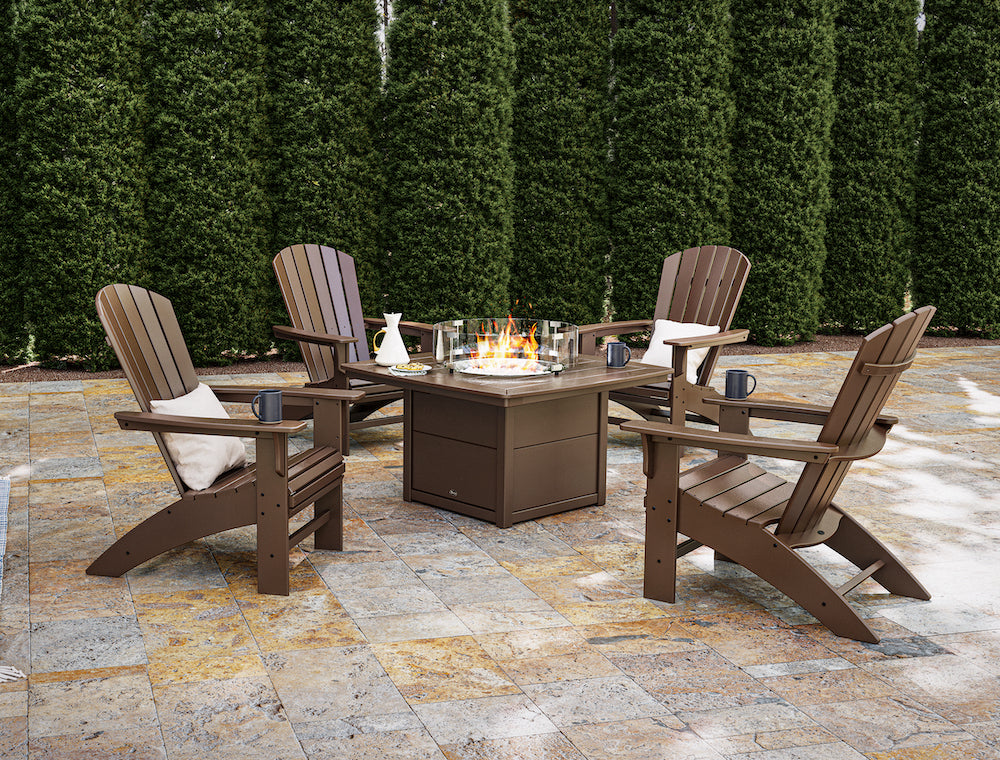 A brown Trex Outdoor Furniture Yacht Club Adirondack 5 Piece Set with Fire Pit Table sits by a row of tall bushes on a patio.