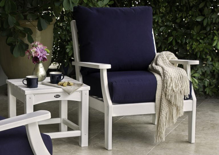 A Trex Outdoor Furniture Yacht Club Deep Seating Chair and Rockport Club 18 Inch Side Table sit in a tiled area by hedges.